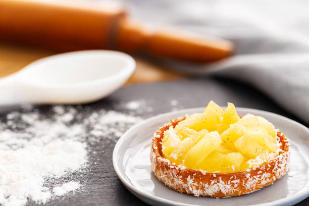Pineapple tart or tartlet with grated coconut in a grey plate, spilled flour, rolling pin and cooking spoon in the background over a slate and napkin. Gray and brown concept. stock photo
