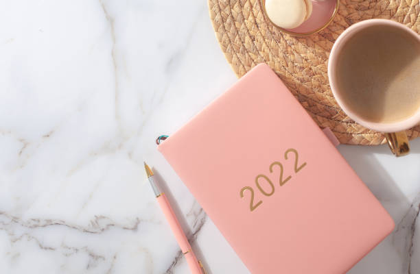 Pink coral colored diary for the year 2022, pen, coffee, macaron cookie on straw woven placemat stock photo