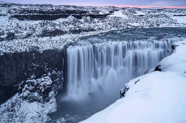 Pink glow in the sky during an early spring sunrise at Dettifoss, Northeast Iceland, where the ground are still covered by blanket of snow. Long exposure captured the water movement of the waterfall. Dettifoss is regarded as the  the most powerful waterfall in Europe which has been featured in Prometheus film. The sediment rich runoffs colours the water greyish white. dettifoss waterfall stock pictures, royalty-free photos & images