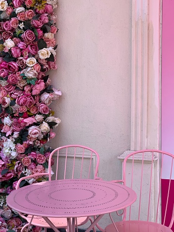 Table et 2 chaises de jardin roses en métal troué . Décoration florales avec des roses sur le mur derrière en guirlande