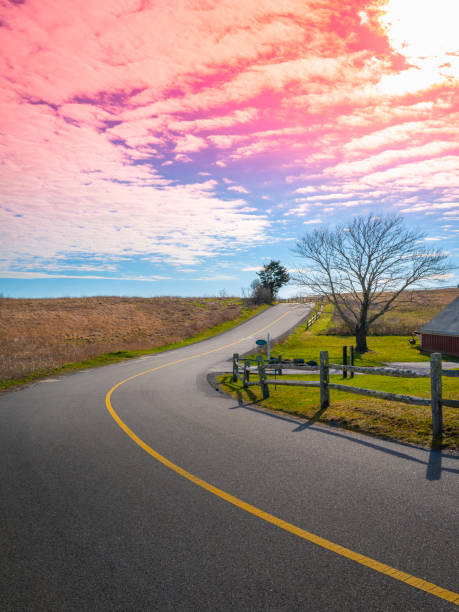 Pink sky and cumulus clouds over the curved uphill asphalt road with wooden fences stock photo