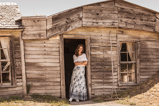 Pioneer woman leaning on wooden prairie homestead in vintage clothing