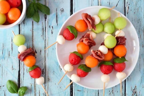 Plate of summer fruit skewers on a rustic blue wood background Plate of delicious summer fruit skewers with melon, cheese and prosciutto on a rustic blue wood background prosciutto stock pictures, royalty-free photos & images