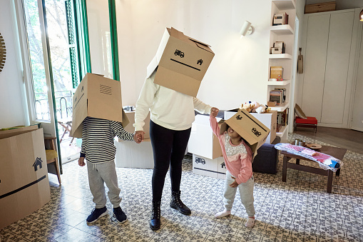 Full length view of Afro-Caribbean mother holding hands of young son and daughter as they take a break from moving to have fun with cardboard boxes.
