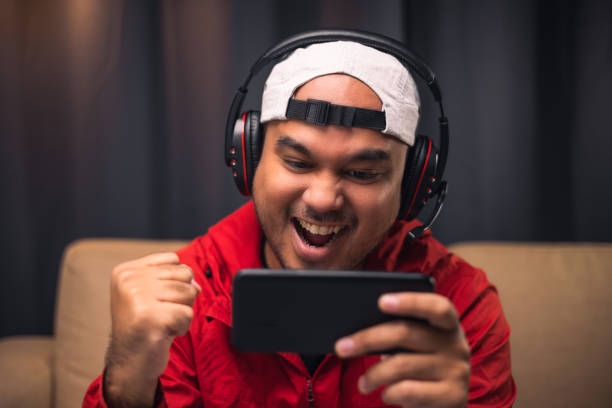 Playing video game on smartphone. Young asian handsome man sitting on sofa holding cellphone in living room. Happiness Streamer Indian man wearing headset playing game online in the darkroom. stock photo