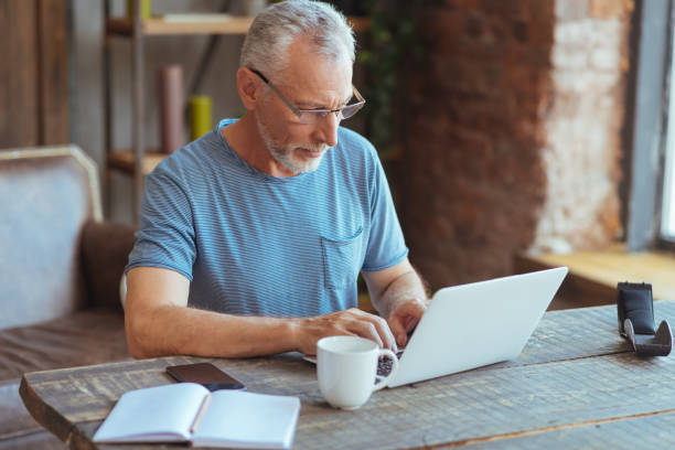 Pleasant retired man being involved in in house work Involved in work. Pleasant handsome retired man sitting at the table and typing while working in house baby boomers stock pictures, royalty-free photos & images