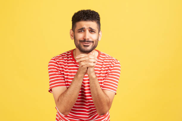 Please, I'm begging! Unhappy bearded man in striped t-shirt keeping hands in prayer looking with imploring expression, sincere asking permission Please, I'm begging! Unhappy bearded man in striped t-shirt keeping hands in prayer looking with imploring expression, sincere asking permission. Indoor studio shot isolated on yellow background prayer request stock pictures, royalty-free photos & images