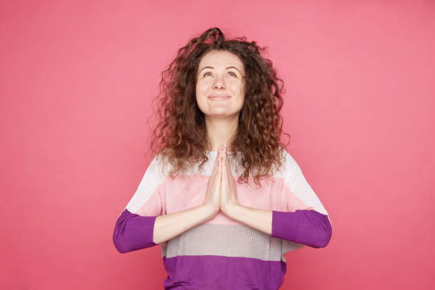 Pleased smiling woman with curly hair Indoor shot of pleased smiling woman with curly hair, keeps palms together in praying gesture, asks for favor, has friendly facial expression, believing God supports and help in difficult situation. prayer request stock pictures, royalty-free photos & images