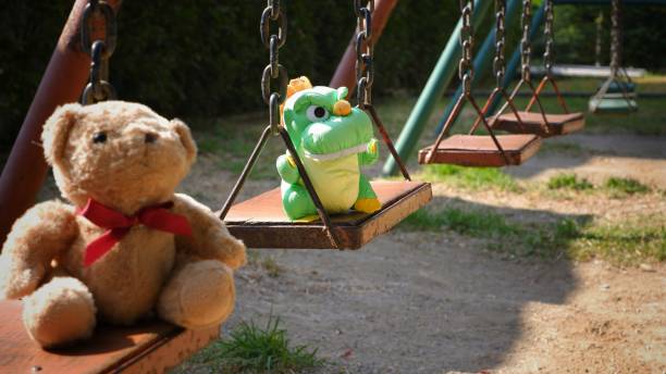 Plush doll on swing set Selective focus at adorable green dinosaur doll with teddy bear sitting on swing set in playground at public park area teddy ray stock pictures, royalty-free photos & images