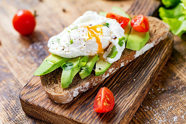 Poached egg and avocado on toast Tasty sandwich with avocado, tomato and poached egg on wooden chopping board, close up, selective focus. Healthy delicious breakfast or lunch poached food stock pictures, royalty-free photos & images