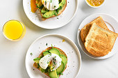 Breakfast with poached eggs on toasted bread with avocado, rukola and herbs and orange juice over white stone background. Top view, flat lay