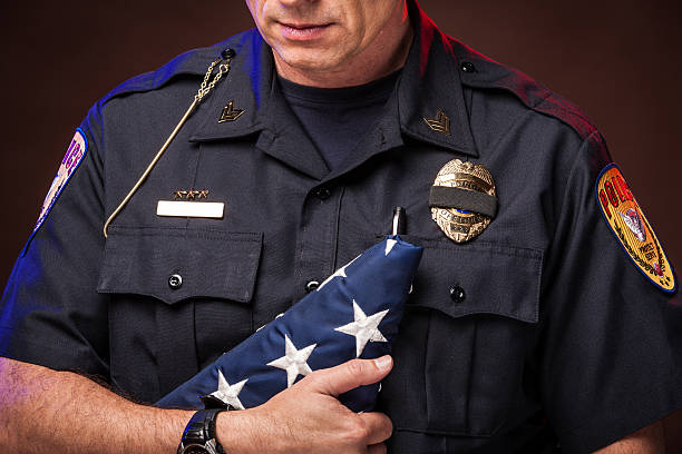 Police Honoring a Slain Officer An authentic police officer in dark, moody lighting. Closeup of a male officer in uniform, holding a folded U.S. flag and looking down in respect, sorrow, and honor for a fallen comrade. A black band covers his badge to signify mourning the death of a fellow officer. mourner stock pictures, royalty-free photos & images