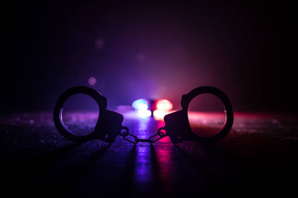Police raid at night and you are under arrest concept. Silhouette of handcuffs with police car on backside. Image with the flashing red and blue police lights at foggy background. stock photo