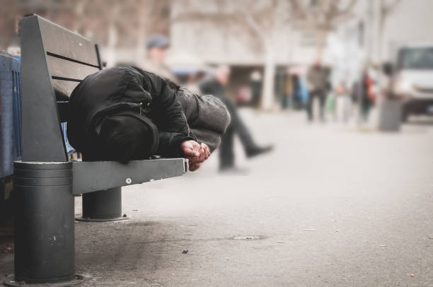 Poor homeless man or refugee sleeping on the wooden bench on the urban street in the city, social documentary concept, selective focus stock photo