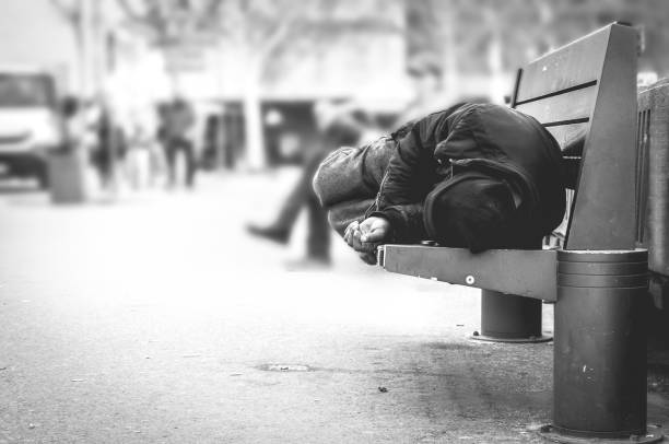 Poor homeless man or refugee sleeping on the wooden bench on the urban street in the city, social documentary concept, selective focus, black and white stock photo