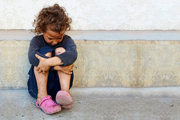 poor, sad little child girl sitting against the concrete wall stock photo
