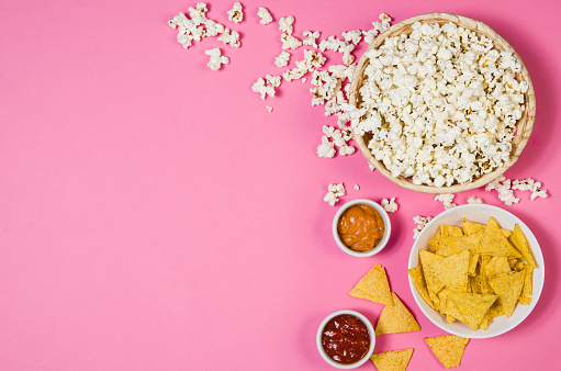 Fresh popcorn, snacks and chips in a bowl isolated on pink background top view. Frame composition with copy space. Movie watching concept