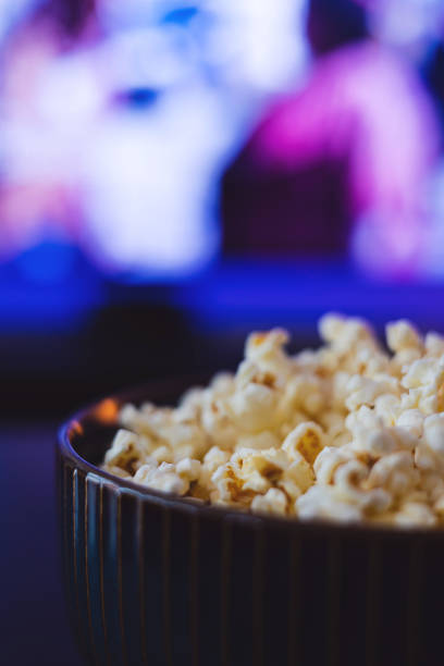 popcorn a bowl with popcorn. in the background a tv and blue dim ambient light. the scene shows home cinema and streaming situation streaming service stock pictures, royalty-free photos & images