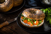 High angle view of a poppy seeds bagel spread with cheese cream and with smoked salmon and arugula topping on a rustic wooden table. The bagel is at the right side of the image and at the top left corner are two defocused bagels on a black plate. Low key DSLR photo taken with Canon EOS 6D Mark II and Canon EF 100 mm f/ 2.8