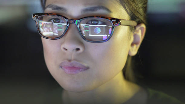 Close up stock image of a young asian woman staring at a computer screen which is reflected in her glasses. The reflection depicts graphs; people & data.