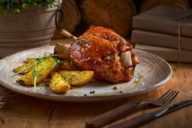 Pork knuckle with baked potatoes on rustic kitchen table at dark wooden background, front view. Pork leg done, German food Pork knuckle with baked potatoes on rustic kitchen table at dark wooden background, front view. Pork leg done, German food. czech culture stock pictures, royalty-free photos & images
