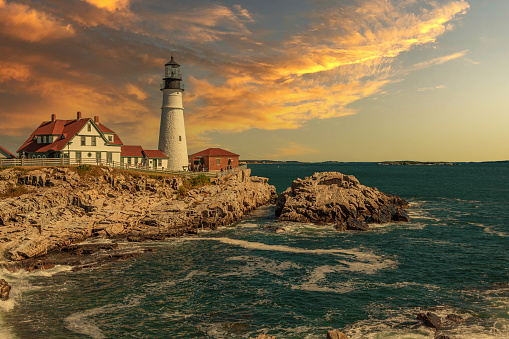 Portland Head Lighthouse, Cape Elizabeth, Maine, lit by the Morning Sun. Rocky Coastline with Cliffs, Ocean Surf and Blue Sky are in the image.