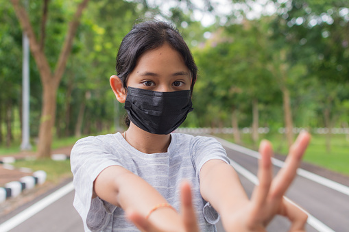 Portrait Asian pretty teen girl in casual dress wears black protective mask posing gesturing with her fingers while standing on the road in public park during COVID-19 epidemic. Healthy lifestyle. Pollution free environment.