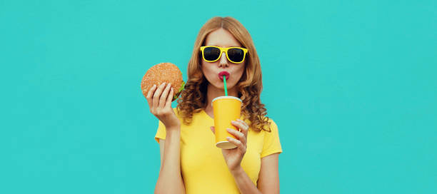 Portrait close up of young woman with fast food, burger and cup of juice wearing a yellow t-shirt, sunglasses on a blue background stock photo