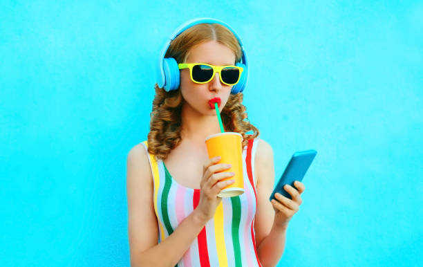 Portrait cool girl drinking fruit juice holding phone listening to music in wireless headphones on colorful blue background stock photo