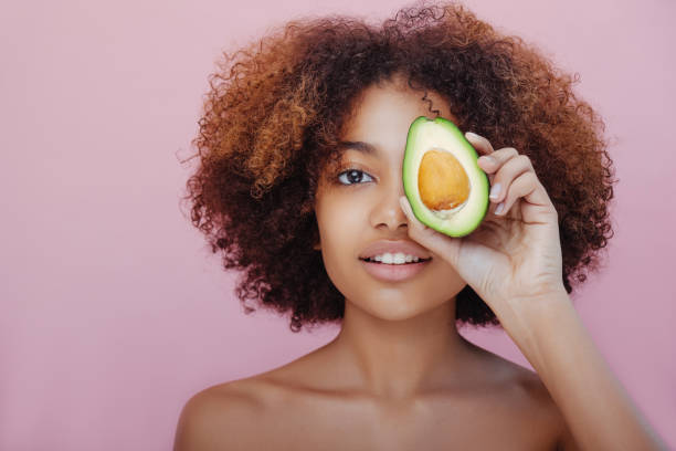 Portrait of a beautiful young black woman closes one eye with half an avocado looks at the camera and smiles stock photo