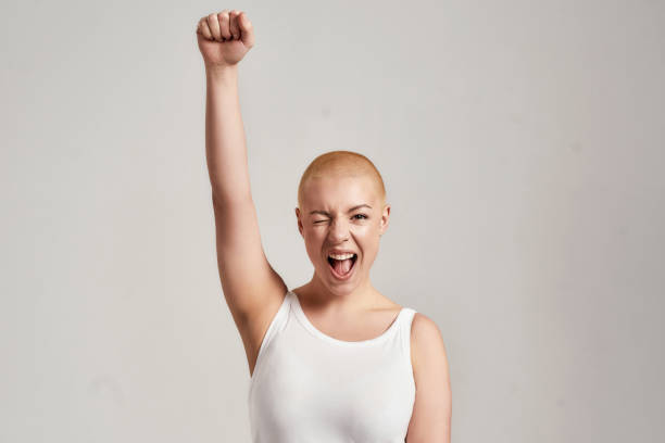portrait of a beautiful young caucasian woman with shaved head wearing white shirt, winking at camera and raising clenched fist, standing isolated over grey background - beleza doentes cancro imagens e fotografias de stock
