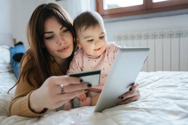 Portrait of a beautiful young mom with a baby girl lying on the bed in a moment of intimacy while buying products online with the tablet and holding her credit card in hand - Millennial with daughter stock photo