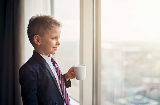 Portrait of a smiling cute blonde boy wearing a suit and a tie. The boys is aged 6 and he's looking at the city holding a cup of coffee or chocolate. The boy is dreaming of becoming a businessman.