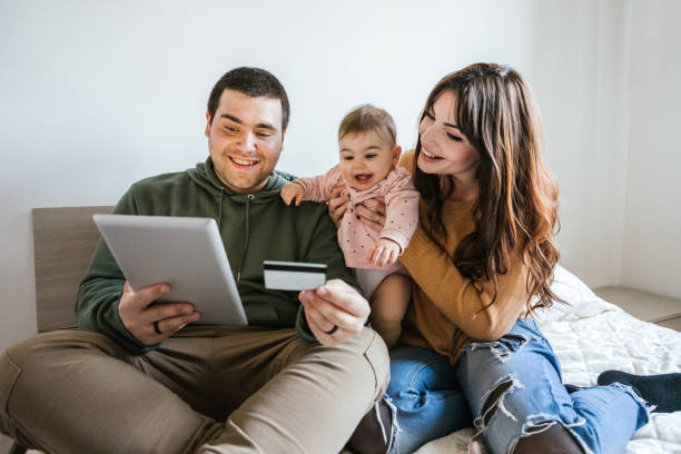 Portrait of a family on the bed looking at the tablet with their baby girl and shopping online with a credit card - Father, mother and little daughter have fun together - Intimacy moment stock photo
