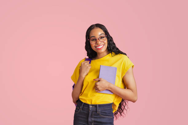 Portrait of a  friendly young female student with book, backpack and glasses, isolated in pink studio copy space stock photo