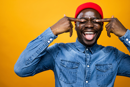 Portrait of a young African American male photo model posing for a photo shoot in a professional model studio with an orange background, wearing denim and a red knit hat, covering his eyes with his fingers and sticking his tongue out
