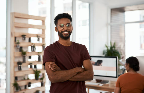 Portrait of a handsome young businessman posing with his arms folded inside an office with his colleagues in the background stock photo