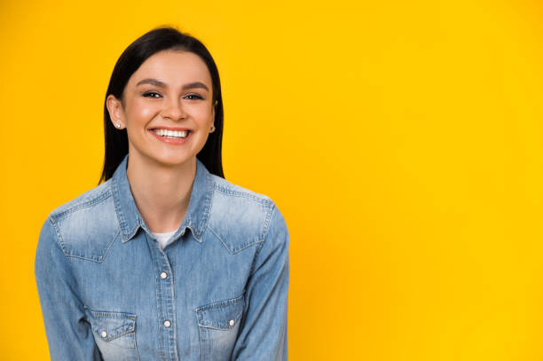 Portrait of a happy smiling caucasian young brunette woman dressed in stylish denim shirt, stands on isolated orange background and looks directly at the camera stock photo