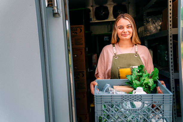Portrait of a Happy Young Caucasian Woman Standing at the Door of a Walk-In Refrigerator Holding a Basket of Delicious, Healthy, Organic Produce at a Local Small Business Farm-to-Table Supplier in Colorado  store food in refrigerator stock pictures, royalty-free photos & images