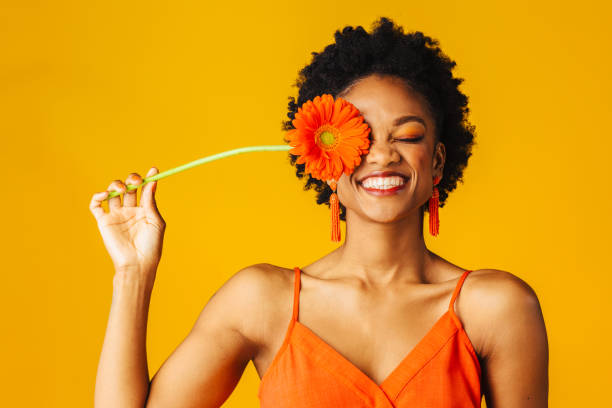Portrait of a happy young woman holding orange Gerbera daisy covering her eye with eyes closed stock photo