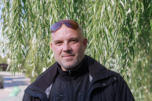 Portrait of a man 40-45 years old European with a good-natured look in the park in autumn in a black jacket against the background of green trees outdoors. Looking into the camera.