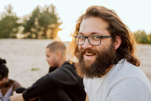 Portrait of a man with glasses, beard and long hair, the guy is wearing a comfortable t-shirt, hipster, smiling, spending time with friends at a bonfire, in the background nature, forests, beach