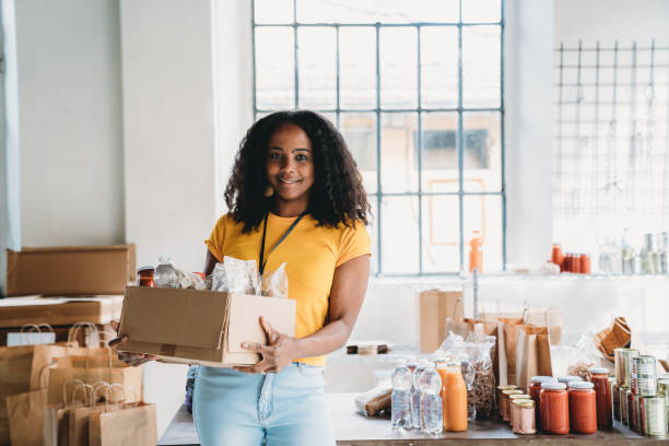 Portrait of a mixed race volunteer woman holding a cardboard box of food and drink at the food bank stock photo