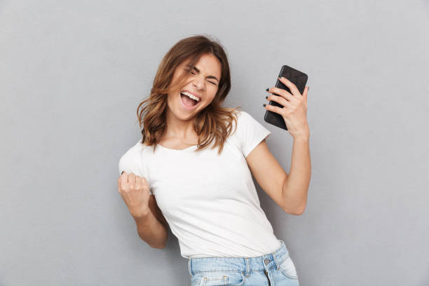 Portrait of a satisfied young woman Portrait of a satisfied young woman holding mobile phone and celebrating over gray background cheerful stock pictures, royalty-free photos & images