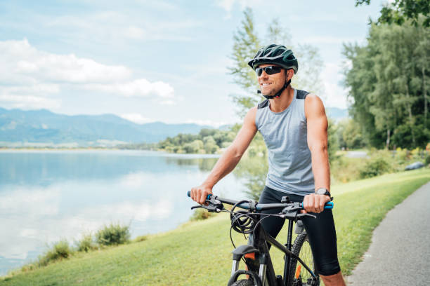 Portrait of a sincerely smiling man dressed in cycling clothes, helmet and sunglasses riding a bicycle on the asphalt out-of-town bicycle path along a mount lake. Active sporty people concept image. stock photo