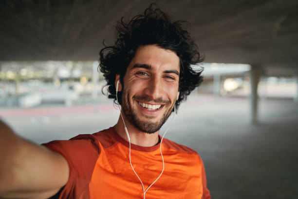 야외에서 셀카를 찍는 귀에 이어폰을 들고 웃는 얼굴의 젊은이의 초상화 - 셀카를 찍고 미소 짓는 카메라를 바라보는 남자의 pov 샷 - selfie 뉴스 사진 이미지