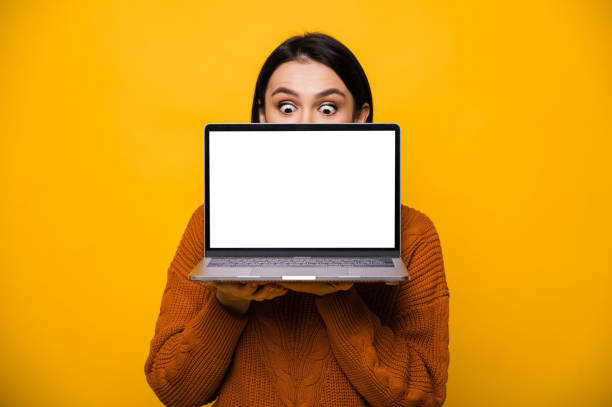 Portrait of a surprised shocked caucasian young woman, hiding behind blank white screen laptop, standing on isolated orange background stock photo