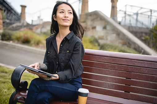 Portrait of a tattooed Asian female in 30's sitting on a bench holding a digital tablet and enjoying her cup of coffee.