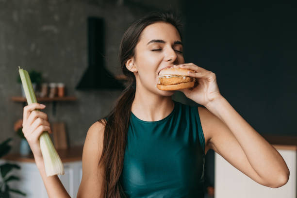Portrait of a woman biting a burger at home, wearing sportwears. Healthy lifestyle concept. Healthy food, diet. Health lifestyle, fitness. Body care. Home concept. stock photo