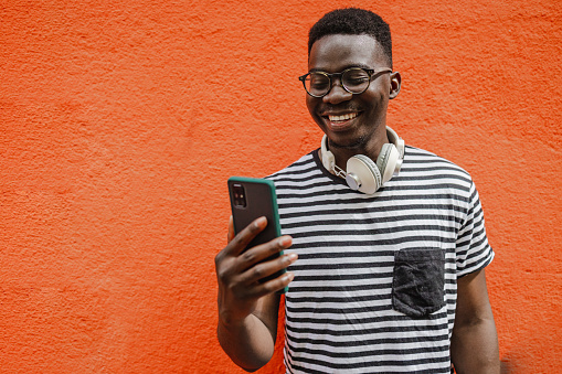 A young African-American man is on the street, he is standing in front of the orange wall and smiling while using a mobile phone